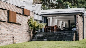 Une maison moderne avec une façade en briques, un espace extérieur aménagé avec une table et des chaises, et une grande plante de type fougère.