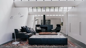 Een moderne woonkamer met een zwarte leren bank, een salontafel en twee zwarte stoelen. Er is een open haard met een zwarte houten plint en een groot abstract schilderij erboven. De kamer heeft witte muren en een groot raam aan het einde.