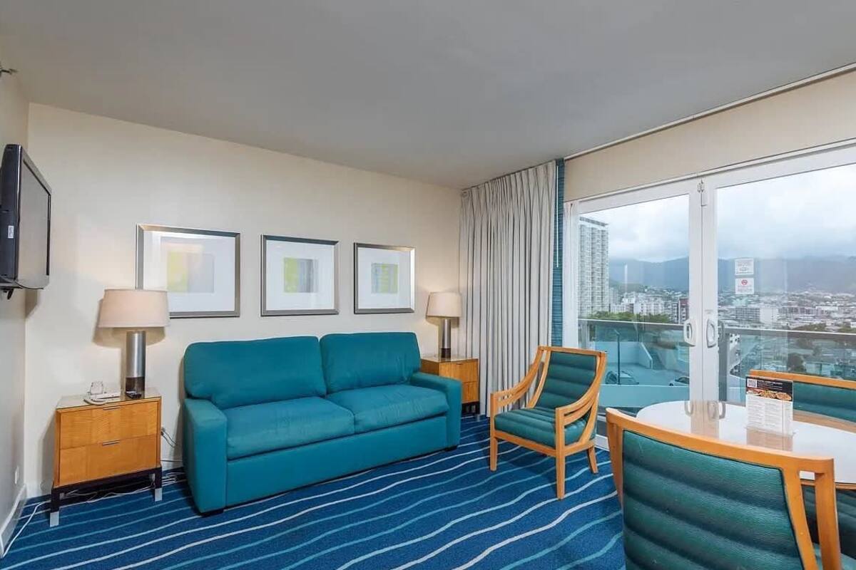 A hotel room with a blue-striped carpet, a teal sofa, a wooden chair, a small table, and a large window offering a city view.