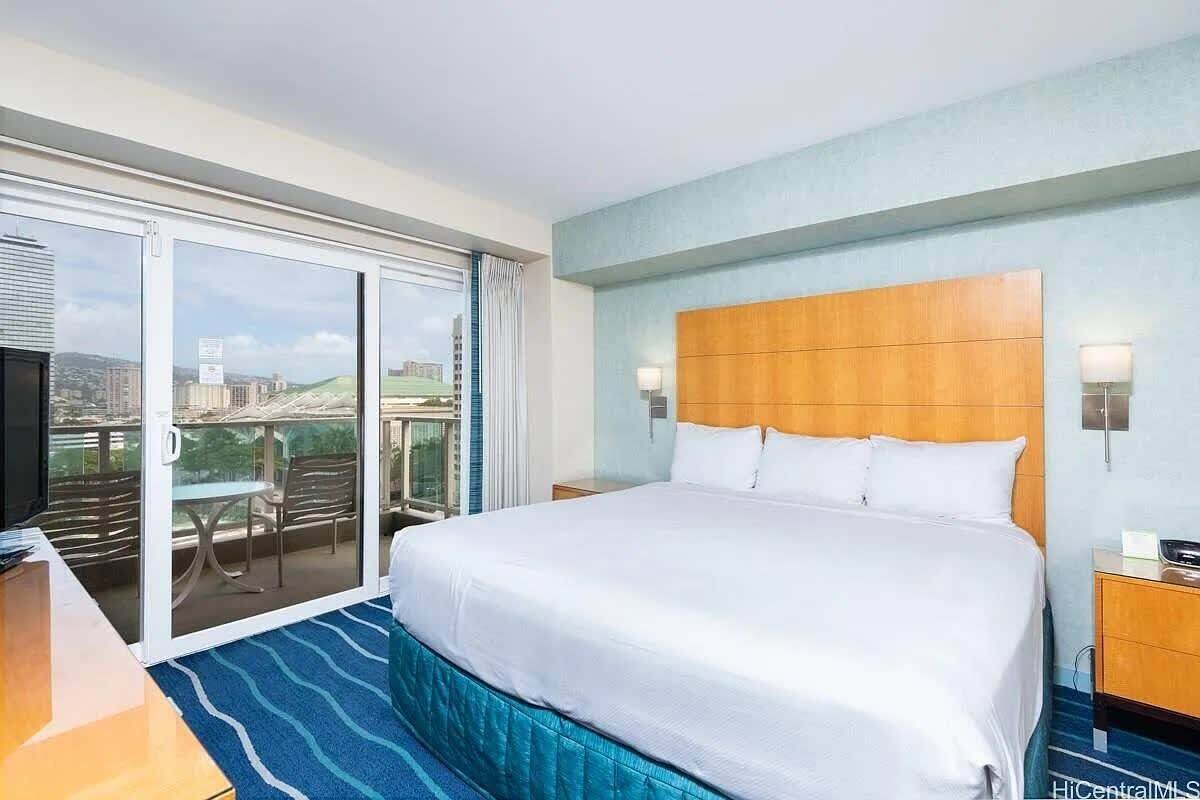 A hotel room with a large bed, a television, a balcony with a table and chairs, and a city view.