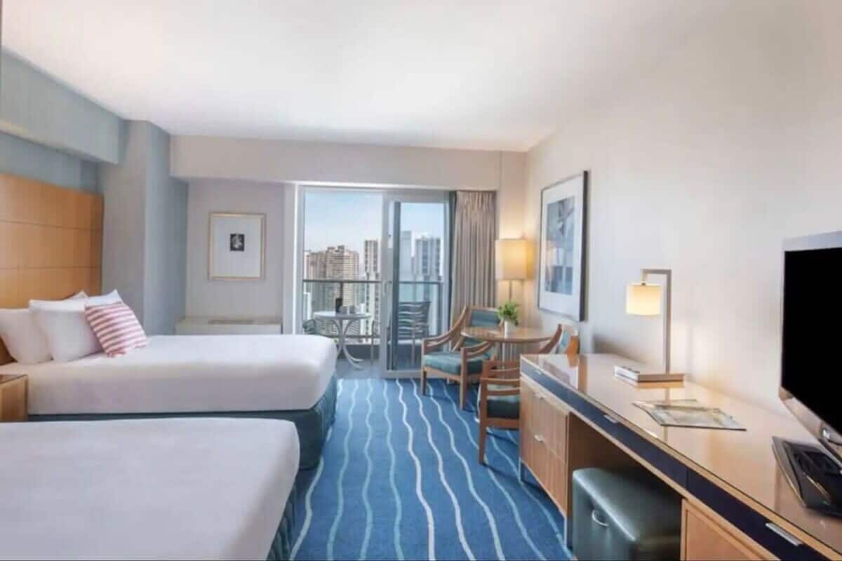 A hotel room with two beds, a desk, a chair, a TV, and a balcony with a city view.