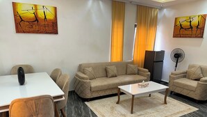 Un salon moderne comprenant un canapé, deux fauteuils, une table basse, une table à manger avec des chaises, et des œuvres d’art accrochées aux murs.