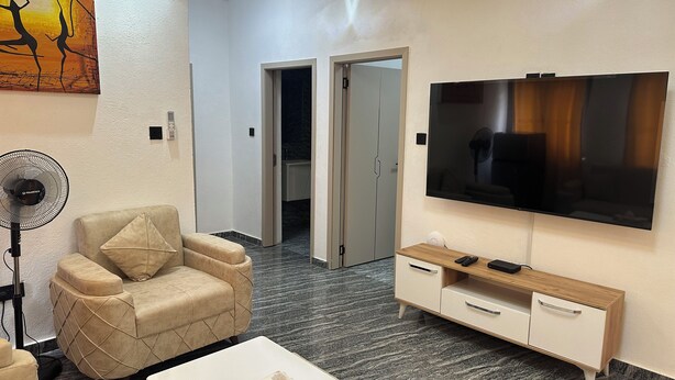 Un salon moderne avec un fauteuil beige, un téléviseur à écran plat, un meuble en bois et un ventilateur.
