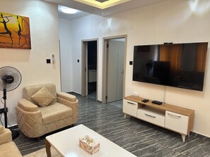 Ein modernes Wohnzimmer mit einem beigefarbenen Sessel, einem Flachbildfernseher, einem Holzschrank und einem Ventilator.