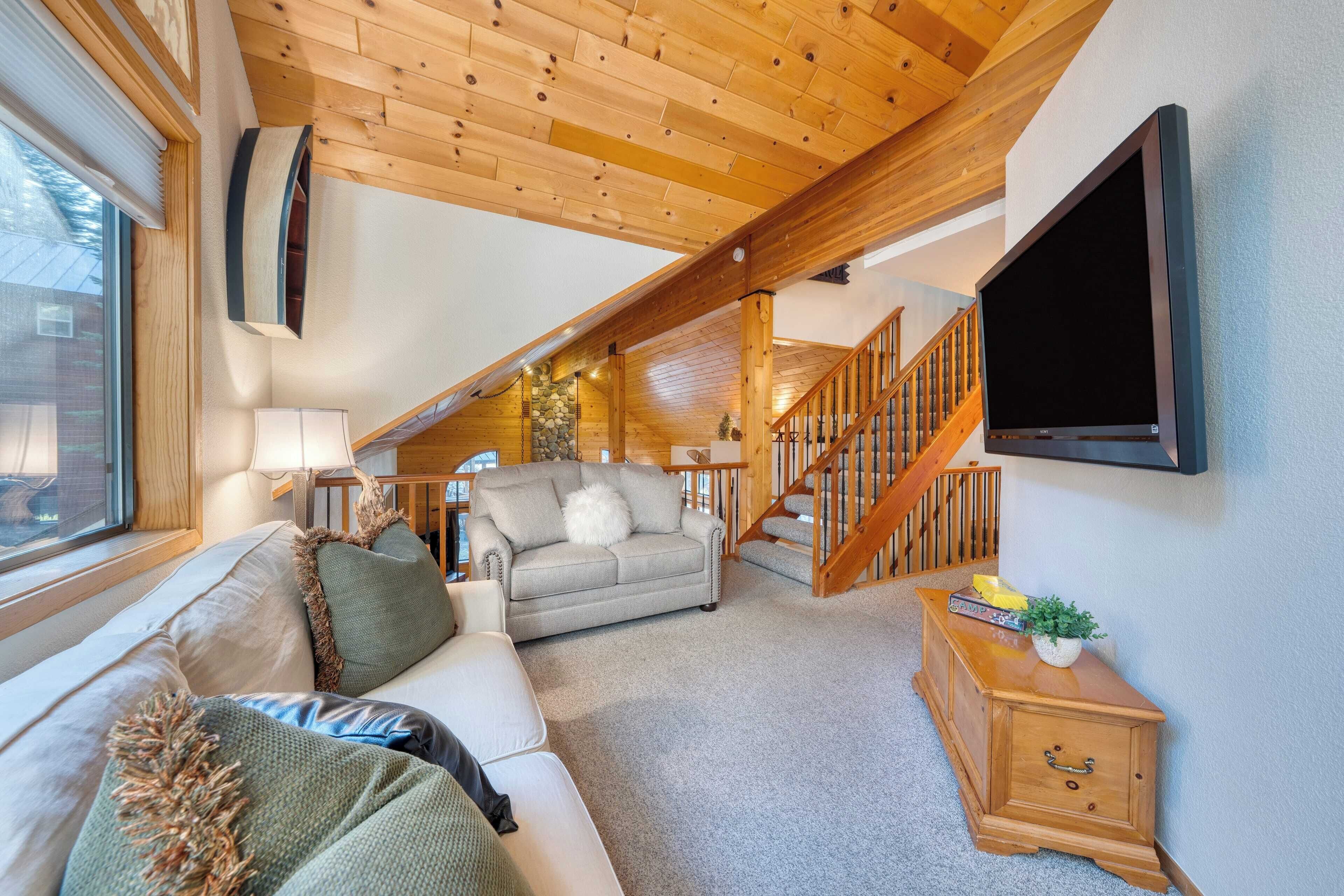 A cozy living room with a wooden ceiling, a sofa, a coffee table, a TV mounted on the wall, and a staircase leading to an upper level.