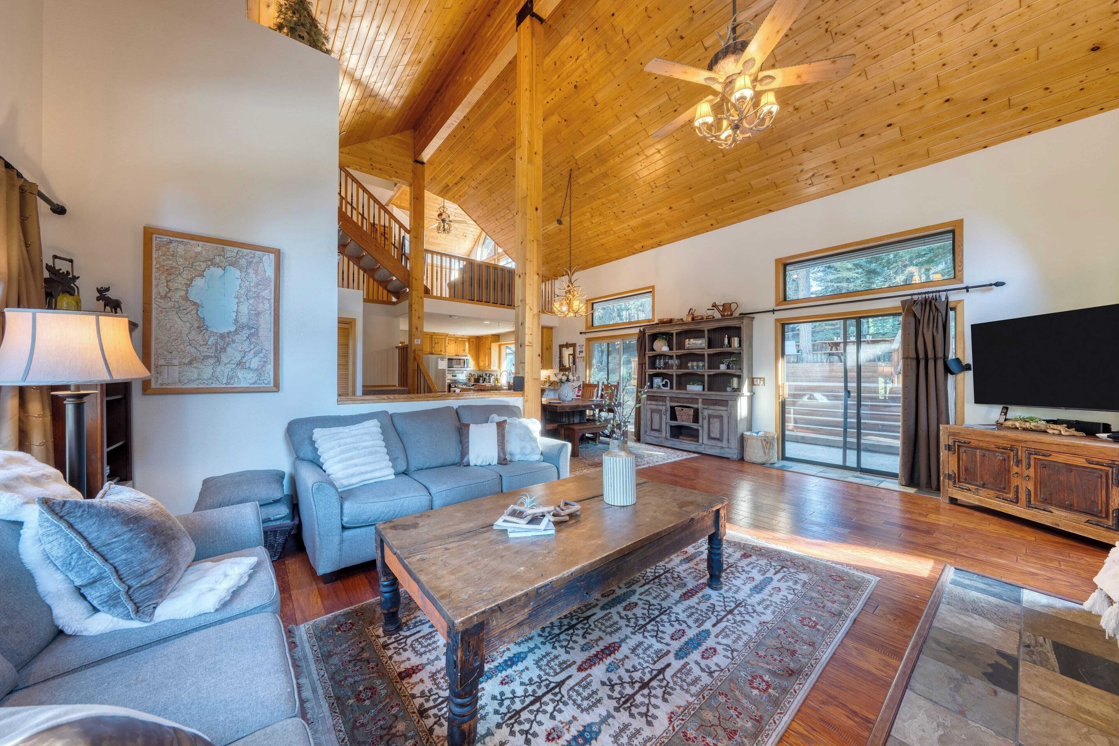 A spacious living room with a high ceiling, a wooden coffee table, a grey sofa, and a large window providing natural light.