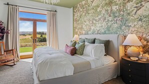 A bedroom with a large bed, a nightstand with a lamp, a view of a garden through a sliding door, and a floral wallpaper. - Wine Country Escape w/ Views + Wine Cellar & Gym Caterina Dusi Vineyard by Avantstay (Paso Robles)