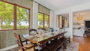 A dining area with a wooden table set for six, large windows with curtains, and a view of trees. - Wine Country Escape w/ Views + Wine Cellar & Gym Caterina Dusi Vineyard by Avantstay (Paso Robles)