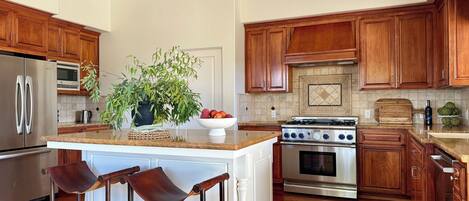 A kitchen with wooden cabinets, a stainless steel refrigerator, a stove, and a large island with a bowl of fruit and a potted plant.
