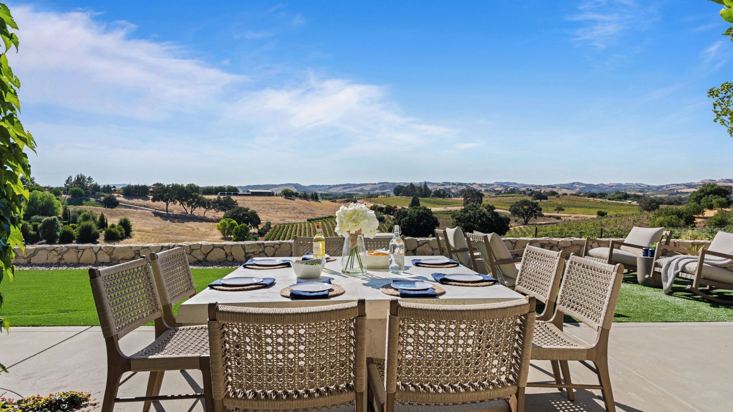 Un espace de restauration en plein air avec une table dressée pour quatre personnes, entourée de chaises en osier, donnant sur un vignoble.