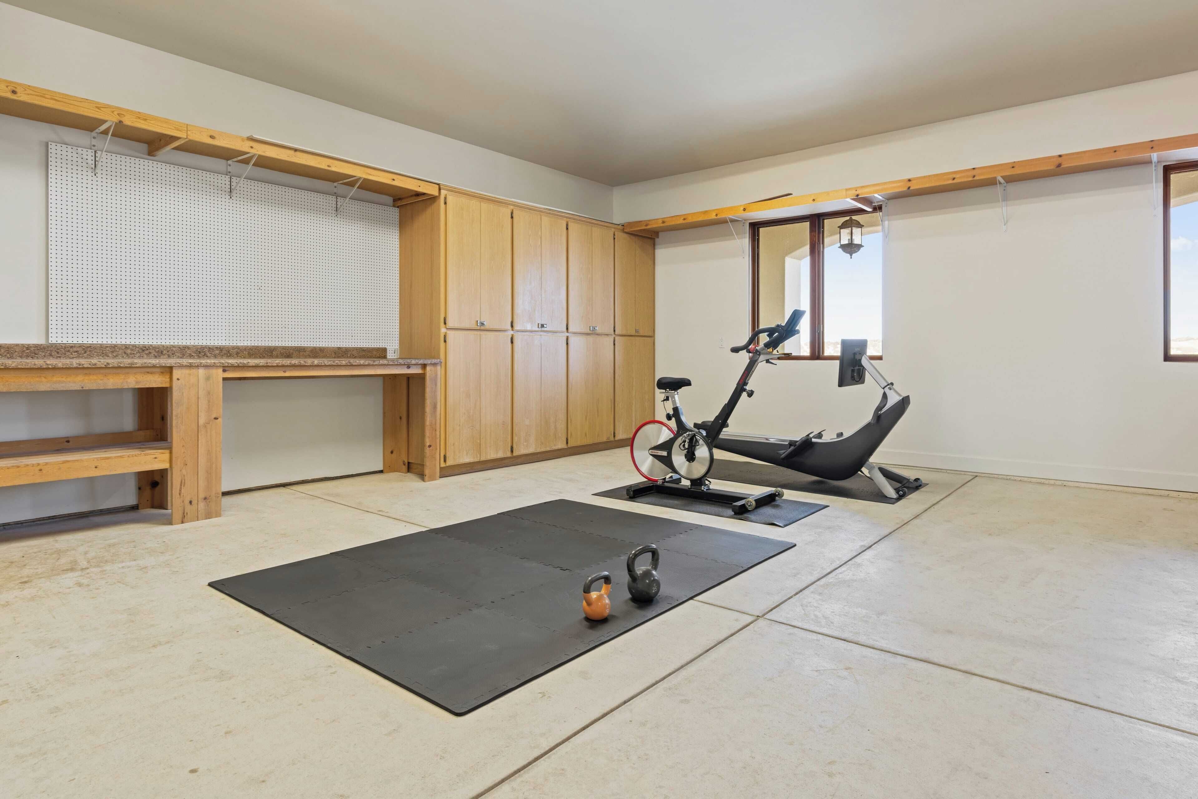 A room with a wooden bench, a black exercise mat, a kettlebell, an exercise bike, and a pegboard wall.