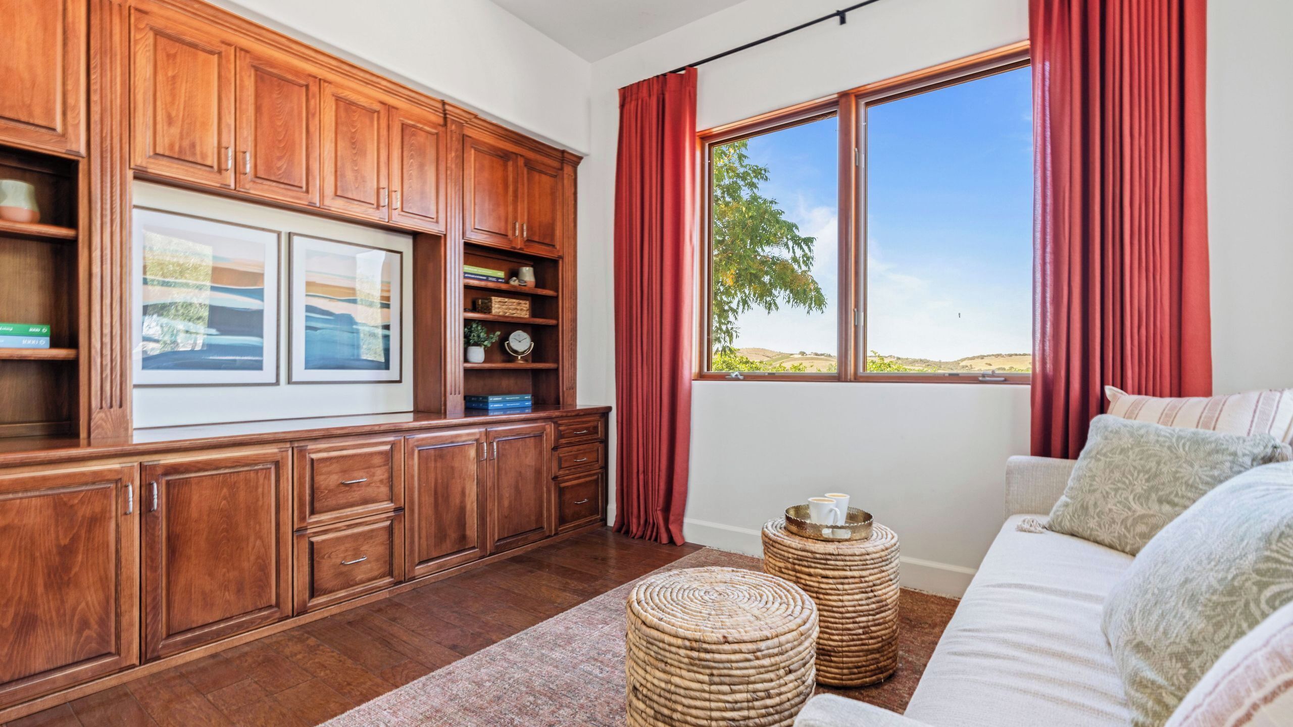 Una habitación con muebles de madera, un ventanal con cortinas, un sofá y otomanas de mimbre.