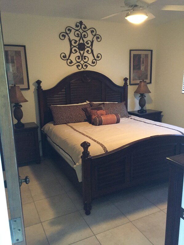 Un dormitorio con cama de madera, almohadas estampadas, dos lámparas de noche, un adorno en la pared y un ventilador de techo.