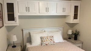 Une chambre à coucher avec un lit, des placards blancs au-dessus, deux tables de chevet avec des lampes et une petite table de nuit avec un tiroir.