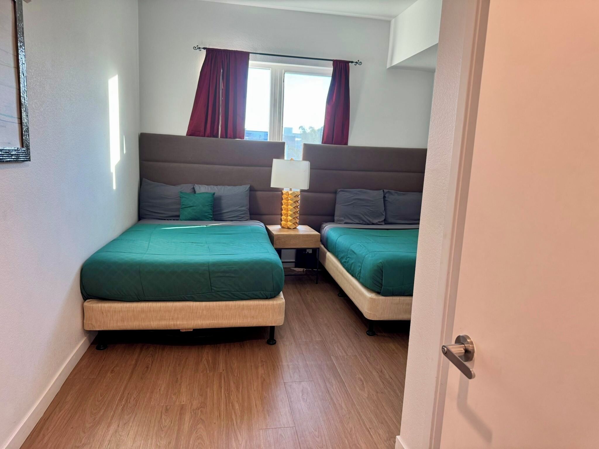 A bedroom with two beds, a window with curtains, a bedside table with a lamp, and a wooden floor.