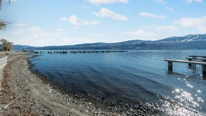 Maisema järven rannalla: puinen laituri ulottuu veteen, rannalla on hiekkaranta ja taustalla kohoavat vuoret.