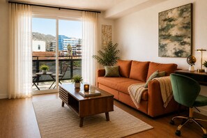 A living room with a sofa, coffee table, and a desk with a chair. There is a plant on the coffee table and a window with curtains.
