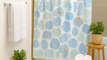 A bathroom with a shower curtain featuring a leaf pattern, a striped rug, two potted plants, and a framed picture on the wall.