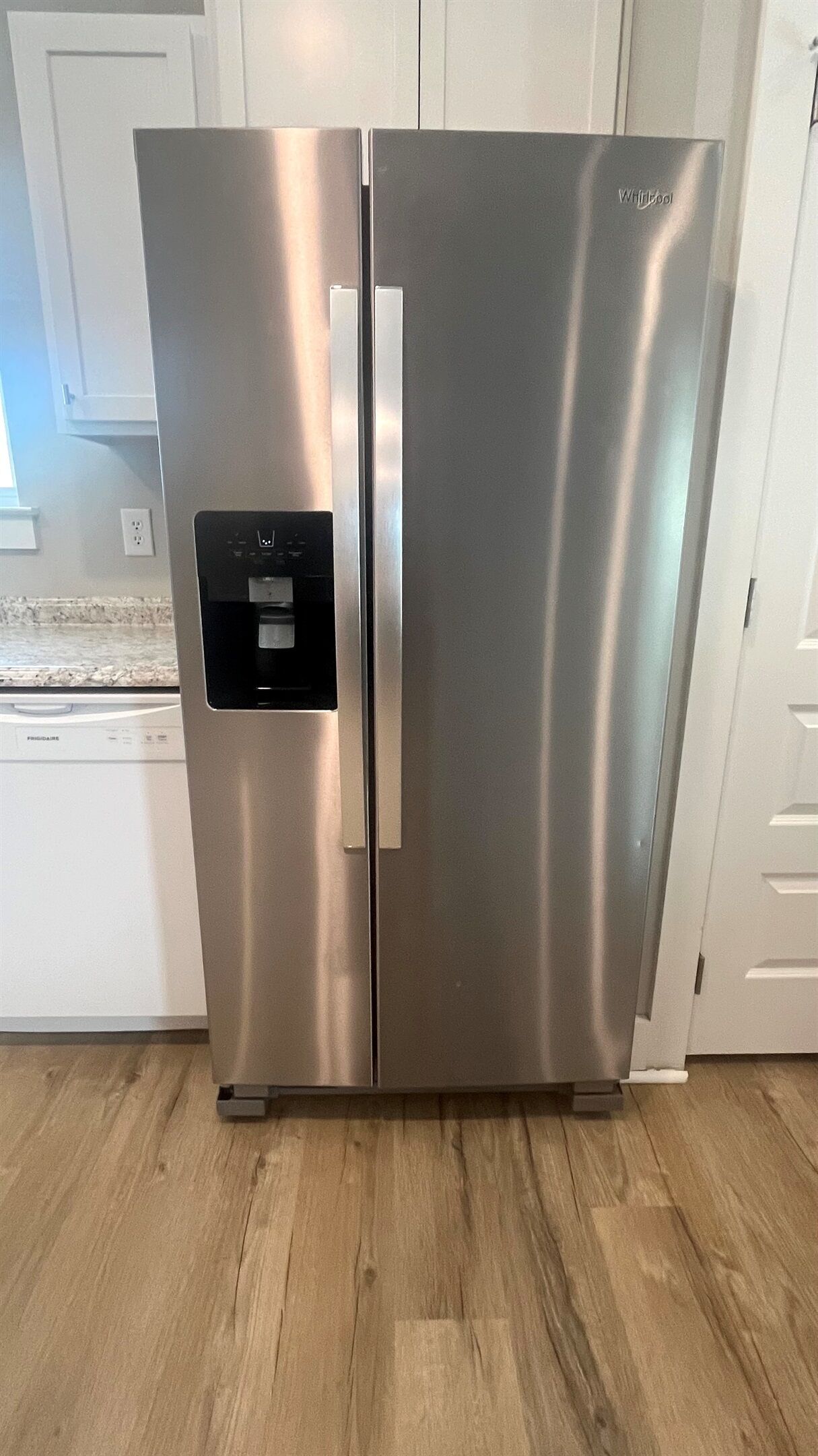 A stainless steel refrigerator with a water dispenser, located in a kitchen with wooden flooring and white cabinetry.