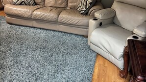 Una sala de estar con un sofá de cuero color beige, un tapete azul con textura y un reloj montado en la pared.