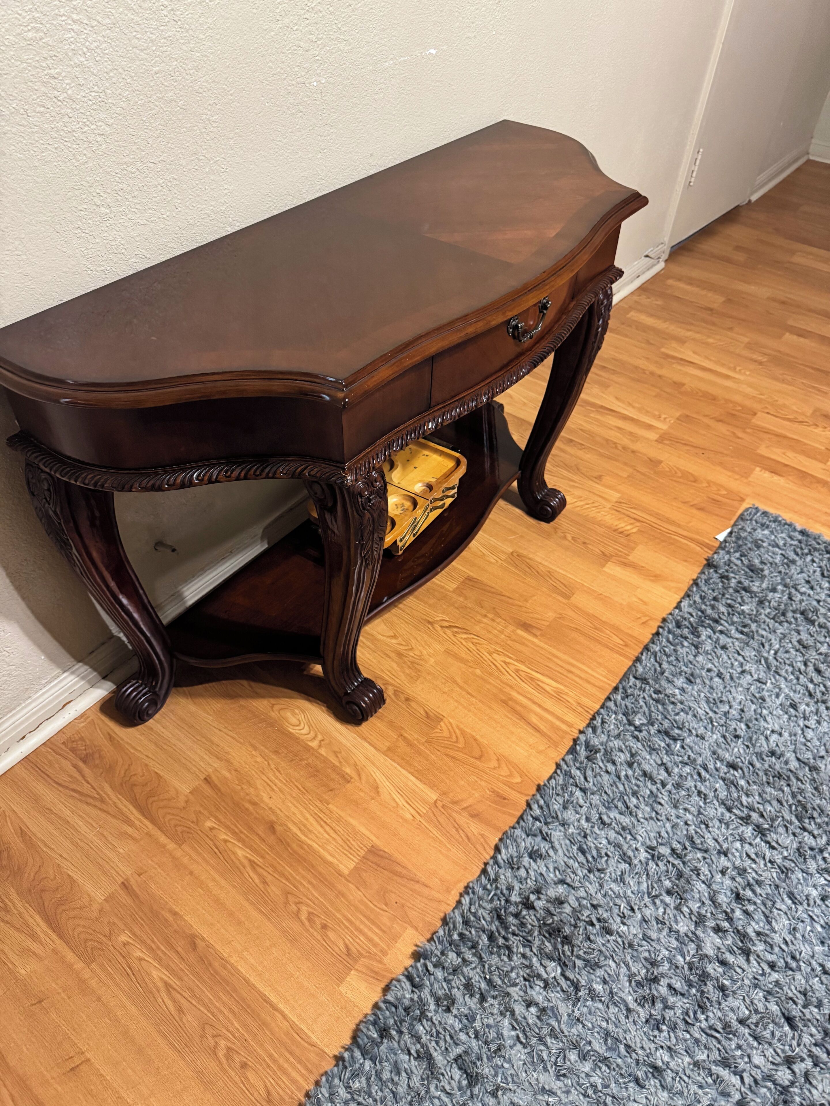 A dark wooden side table with a drawer and a small shelf, placed on a wooden floor next to a white wall and a gray rug.
