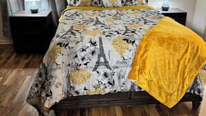 Una cama bien hecha con una colcha con motivos de París, dos lámparas de noche y un cuadro en la pared sobre la cama.