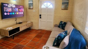 Un saló amb televisió de pantalla plana penjada a la paret, un moble de fusta, un sofà beix amb coixins estampats i un paviment de rajola.