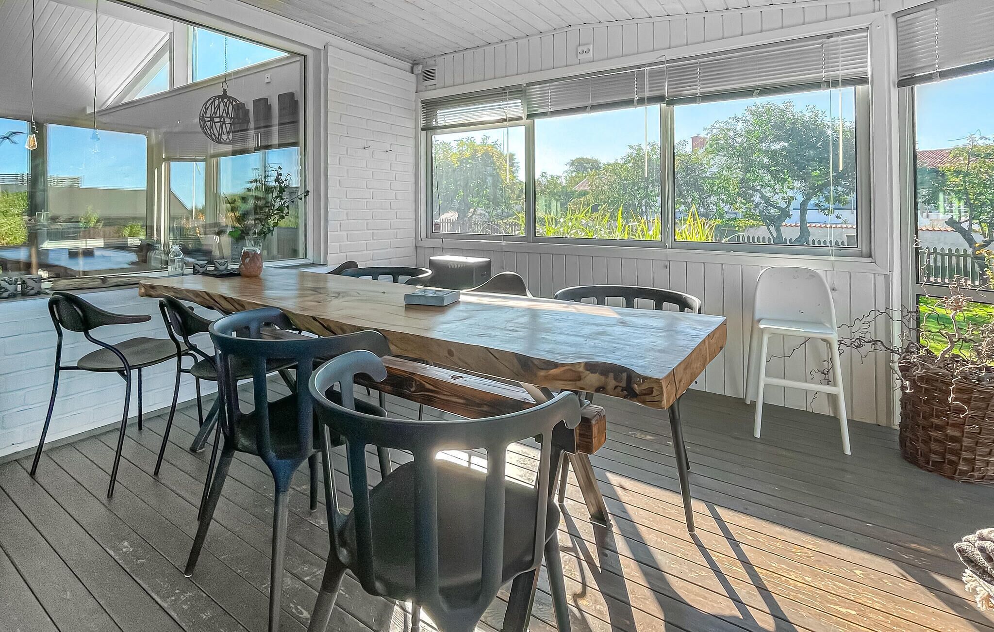 Un vaste espace de salle à manger avec une longue table en bois et des chaises modernes, de grandes fenêtres offrant une vue sur la verdure et une terrasse qui s’étend à l’extérieur.