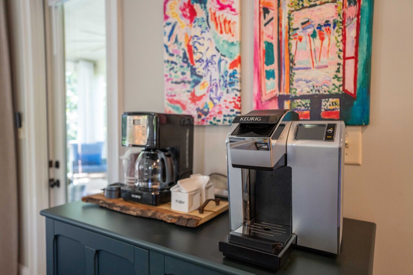 Een koffiehoek met een Keurig koffiezetapparaat en een koffiekan op een houten plank, met kleurrijke kunstwerken aan de muur.