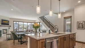 A modern kitchen with a central island, wooden cabinets, pendant lighting, a staircase, and a dining area with a view. - Brand-New 4BR Townhome | Private Hot Tub (Fraser)