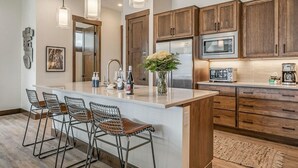 A kitchen with wooden cabinets, a stainless steel refrigerator, a microwave, and a coffee maker. There are four bar stools and a vase with flowers on the island. - Brand-New 4BR Townhome | Private Hot Tub (Fraser)