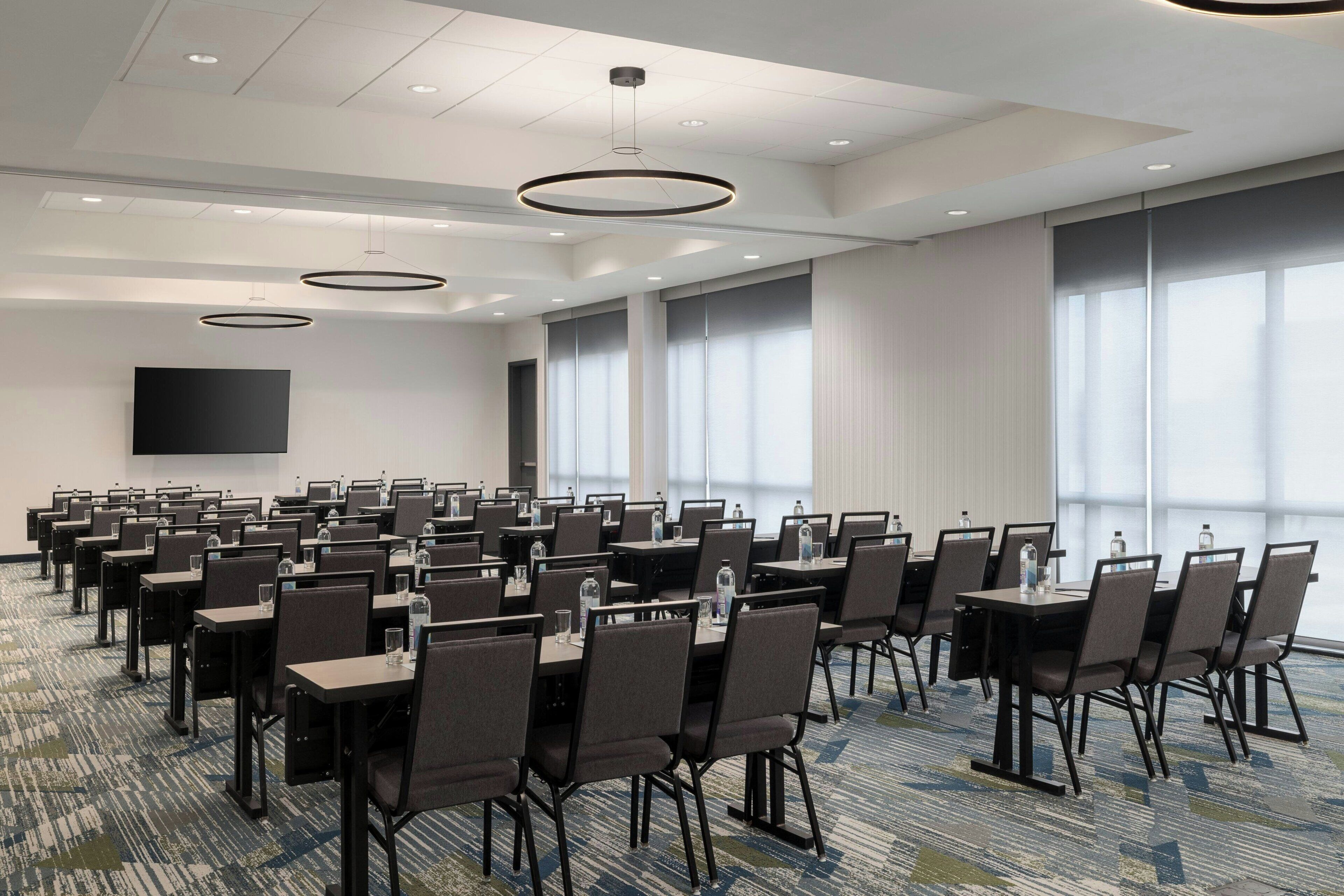 A conference room with rows of tables and chairs, large windows, and a flat-screen TV on the wall.