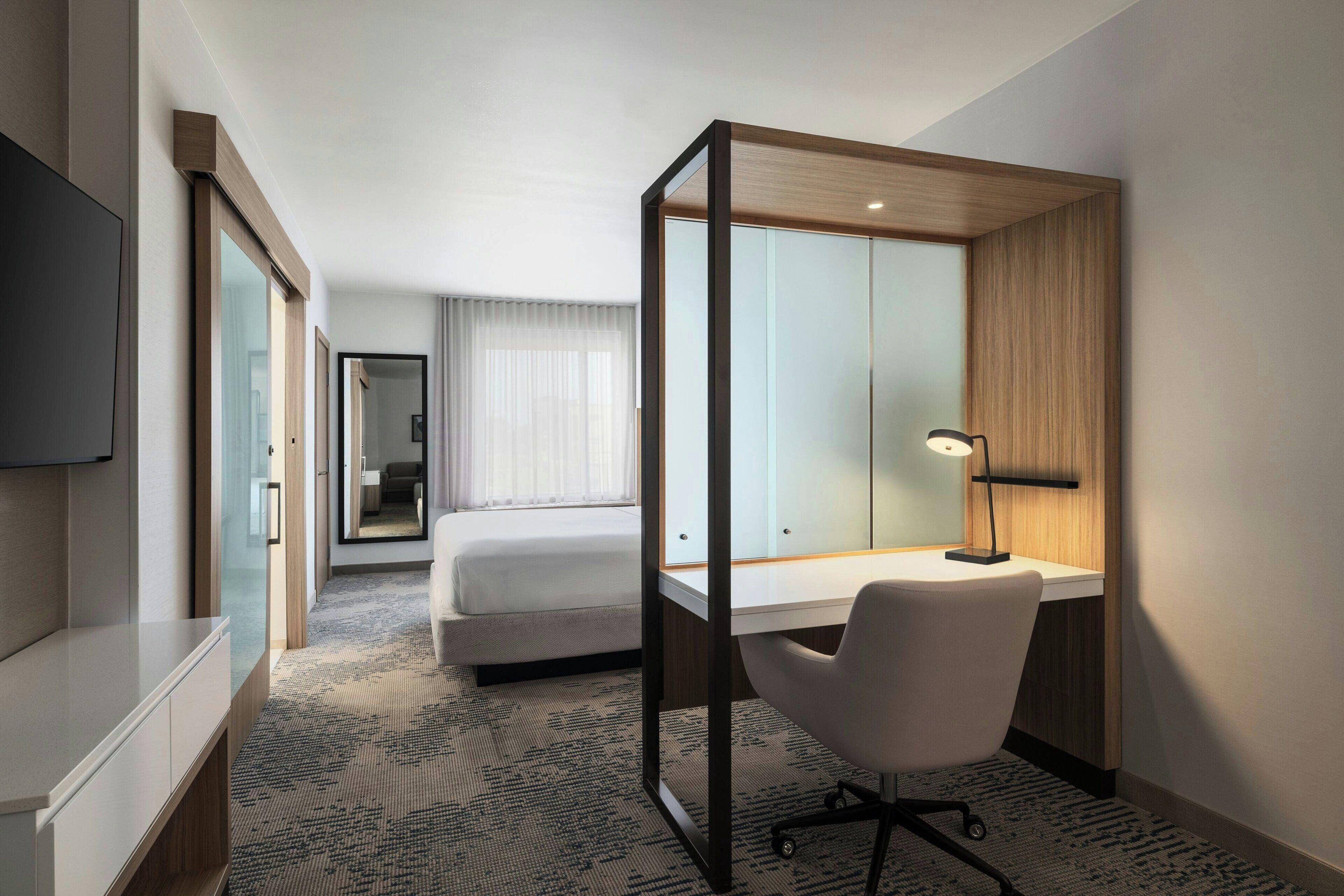 A hotel room with a bed, a desk, a chair, a TV, and a mirror.