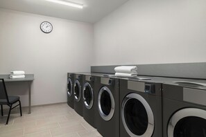 A laundry room with four front-loading washing machines, a wall-mounted clock, and a chair. - Springhill Suites By Marriott Abilene (Abilene)