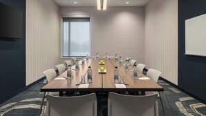 Uma sala de conferências com uma mesa comprida, cadeiras brancas, garrafas de água e uma tela de projetor.