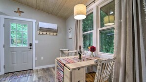 A room with a wooden table, a chair, a window with curtains, a hanging light fixture, and an air conditioning unit on the wall. - Cozy Mountainside Studio Nestled in the Trees (Sylva)