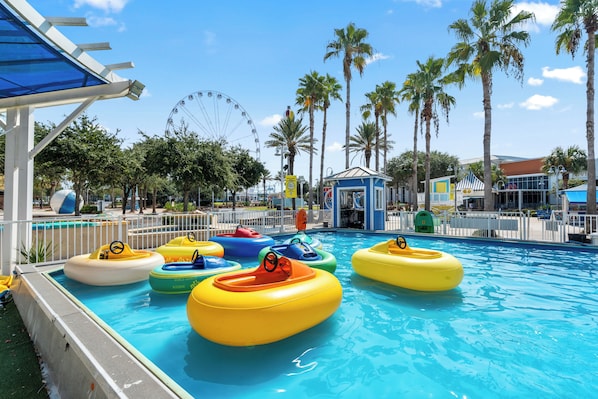 A swimming pool with inflatable rings, palm trees, and a Ferris wheel in the background.