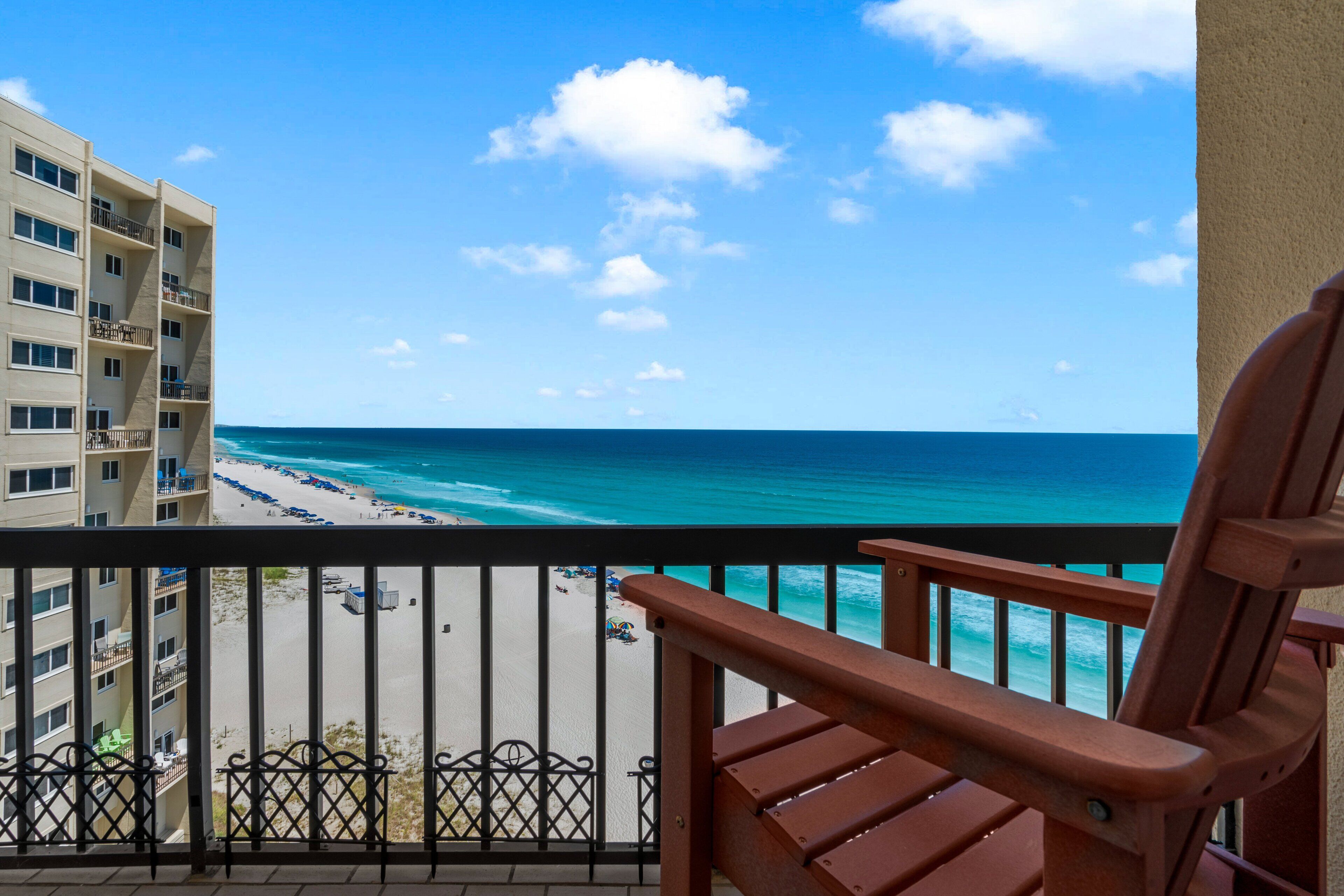 Ein Balkon mit Blick auf Strand und Meer, ein Holzstuhl und ein mehrstöckiges Gebäude.