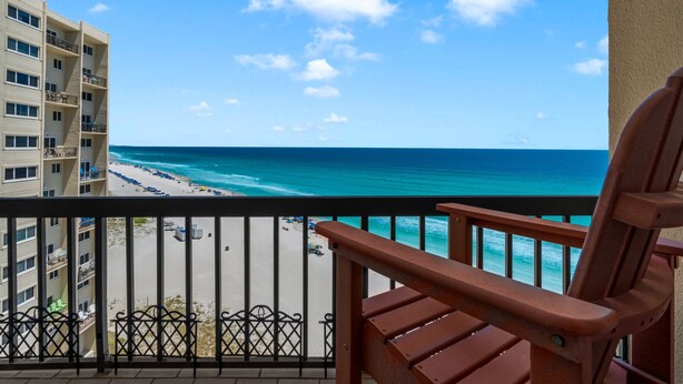 Un balcone con vista su una spiaggia e l'oceano, una sedia di legno e un edificio a più piani.