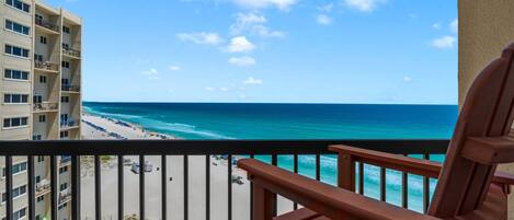 A balcony with a view of a beach and ocean, a wooden chair, and a building with multiple floors.