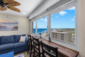A room with a view of the ocean, a sofa, a dining table with chairs, and a ceiling fan.