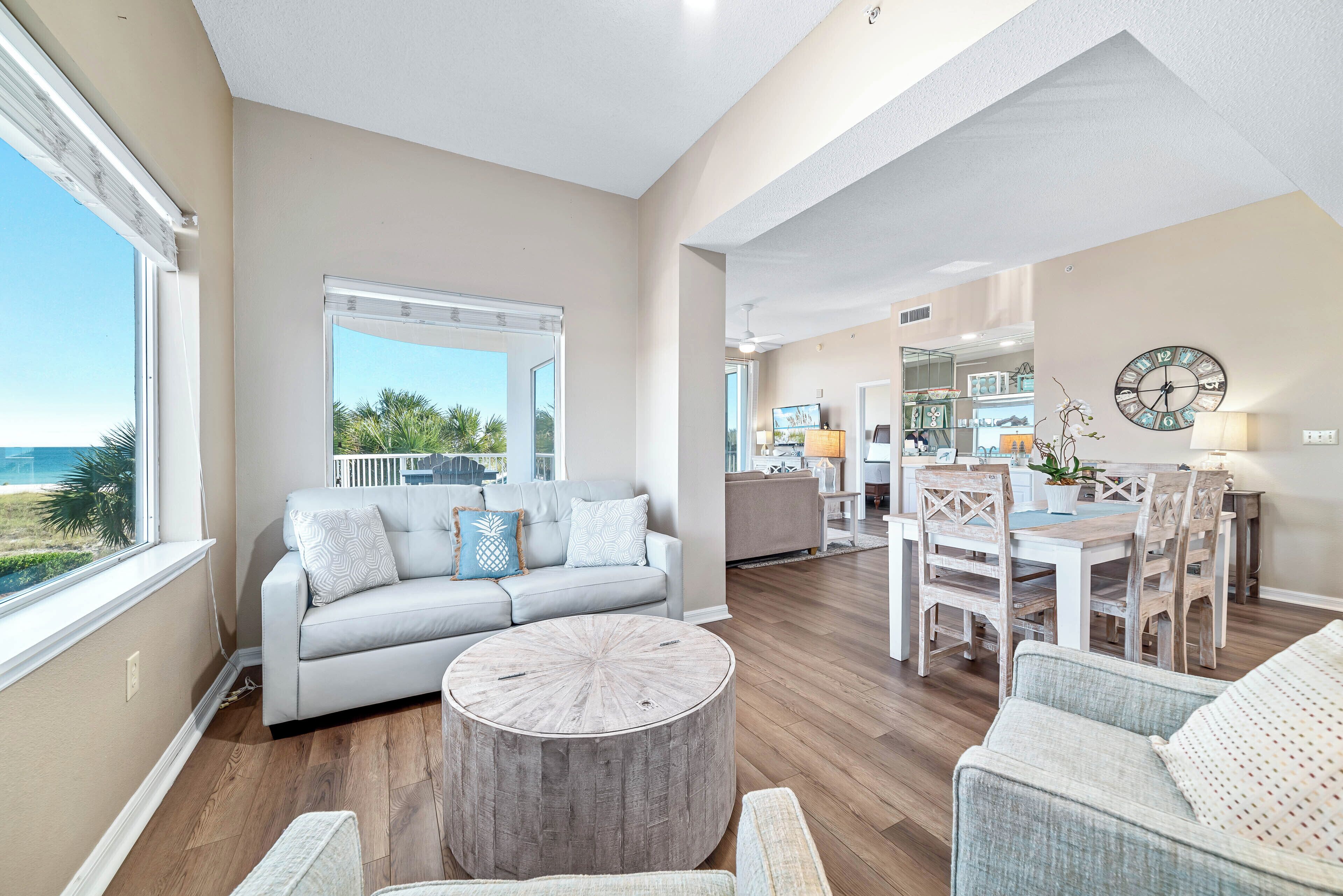 A spacious living area with a large window, a gray sofa, a wooden coffee table, and a dining area with a white table and chairs.