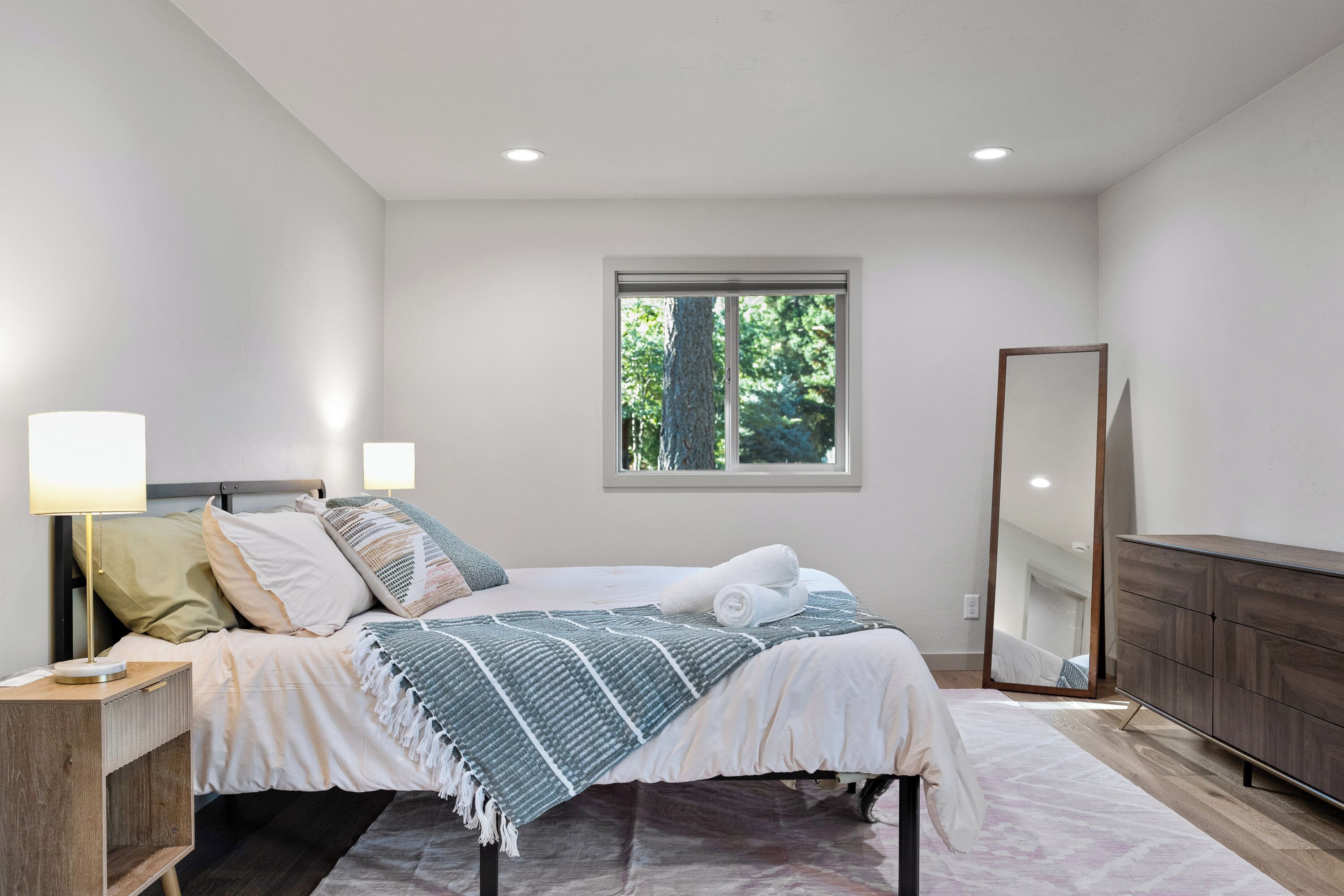 A modern bedroom with a bed, bedside tables, a mirror, and a window with a view of trees.