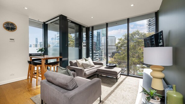 A modern living room with a sofa, a dining table with chairs, a TV, and large windows offering a city view.