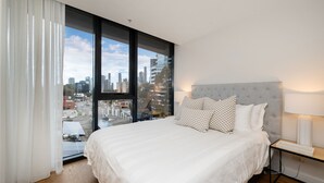 A modern bedroom with a large bed, a grey upholstered headboard, and a view of a cityscape through floor-to-ceiling windows.
