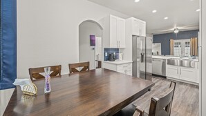A modern kitchen with white cabinets, a stainless steel refrigerator, and a dining area with a wooden table and chairs. - Welcome to The Chelsea Inn! This cozy, modern 3BR/2BA home is designed for all! (Tampa)