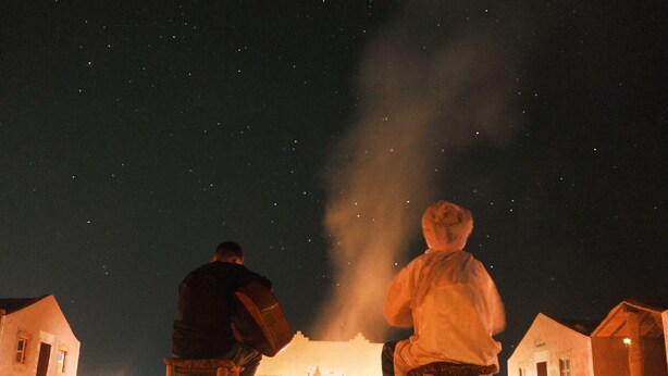 Two people sitting on stools under a night sky with stars, in front of a building with a visible chimney emitting smoke. - Charming Berber camp in Mhamid Desert  (Zagora)