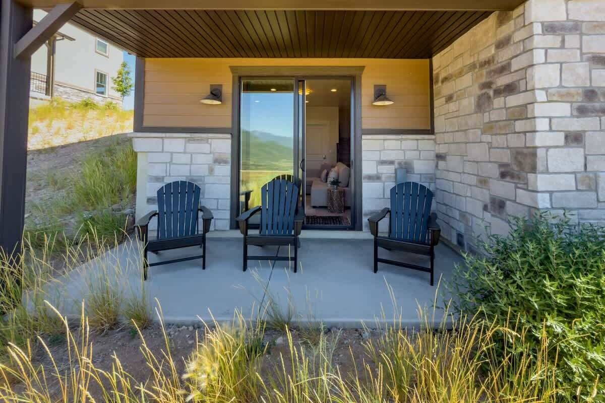 A covered outdoor seating area with three chairs, a stone wall, and a view of a building in the background.