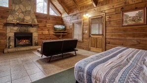 A log cabin interior with a stone fireplace, a television, a sofa, and a bed. - Tranquility - Hot-Tub, Fireplace (Pigeon Forge)