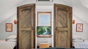 Un dormitorio con una televisión grande montada en una pared de madera, dos camas con ropa de cama blanca y un pequeño cofre de madera.